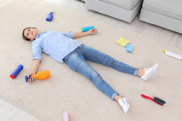 Exhausted woman lying on carpet with stain removers, brushes, cloths, and other cleaning supplies strewn around her